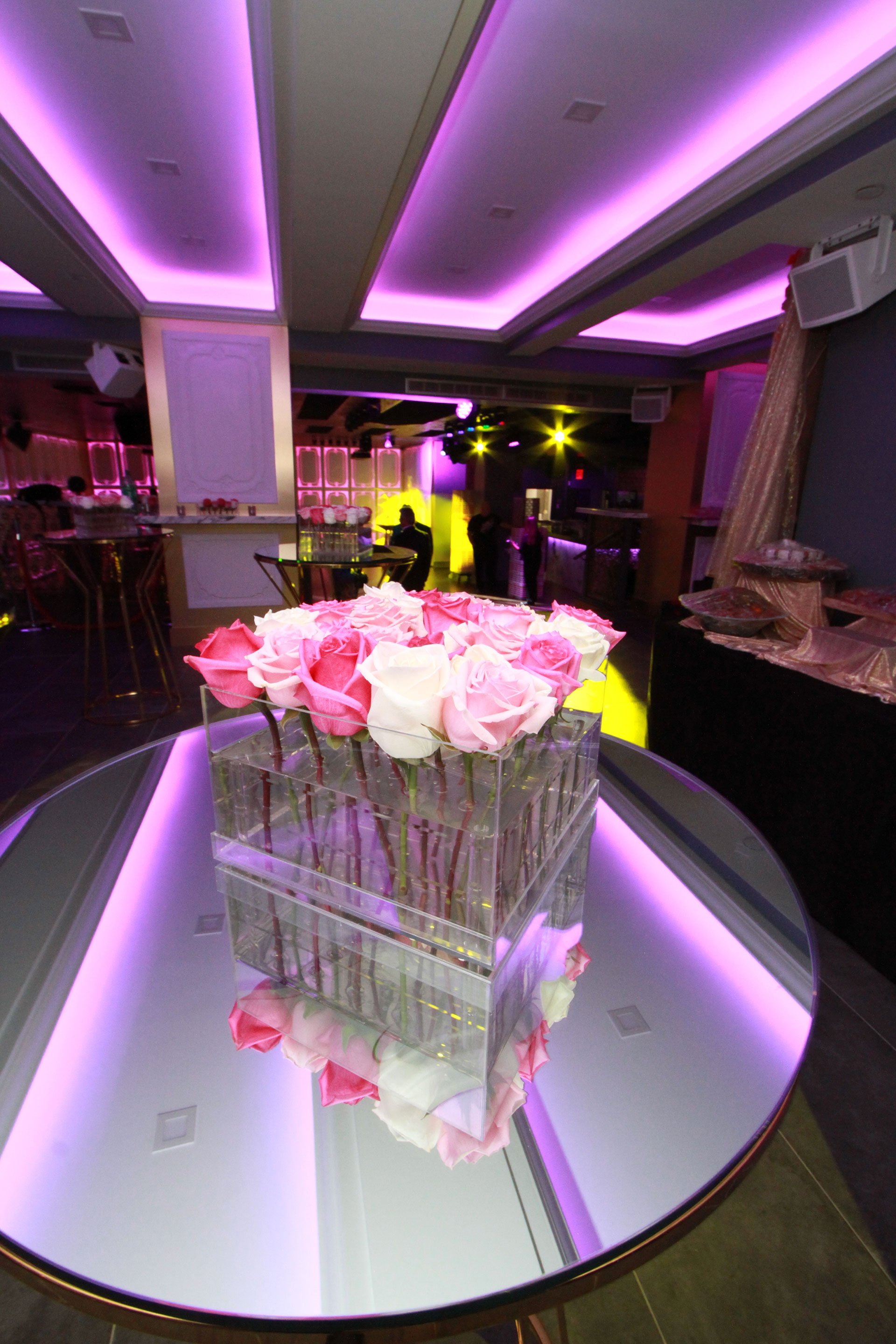 An elegant event space with a purple lighting scheme, featuring a round dining table adorned with a clear vase containing pink roses.